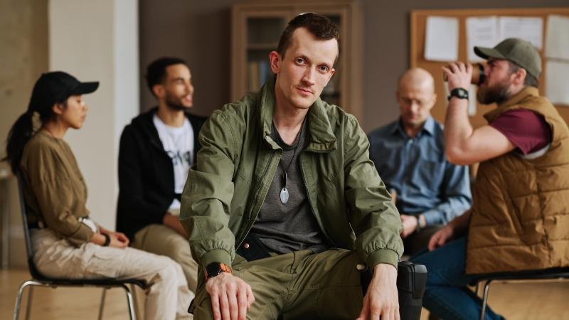 Man in a bomber jacket sitting in front of other people in a support group circle